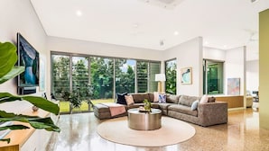 A spacious living room with a large sectional sofa, a round coffee table, and a view of the outdoors through floor-to-ceiling windows. - ZENLUX - 5BR Holiday Home with Pool (Fannie Bay)