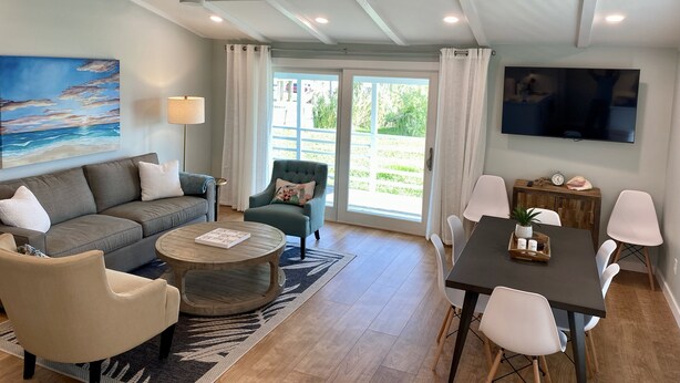 A modern living room with a sofa, armchairs, a coffee table, and a dining area with a table and chairs. There is a painting on the wall, a ceiling fan, and a TV mounted on the wall.