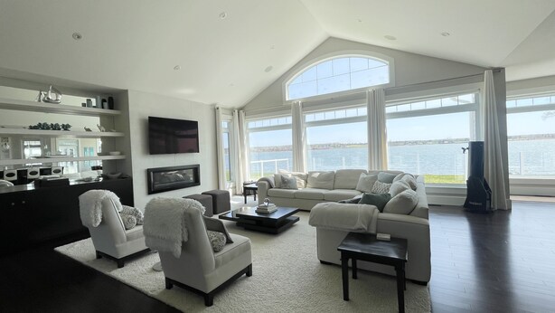 A spacious living room with a large window offering a view of the water, a flat-screen TV mounted on the wall, and a modern kitchen area with built-in appliances. - Executive Water Front Home With Steps To Water, 15 Mins to Downtown (Cornwall)