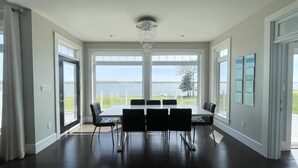 A spacious dining area with a large table and chairs, large windows with a view of the water, and a modern chandelier. - Executive Water Front Home With Steps To Water, 15 Mins to Downtown (Cornwall)