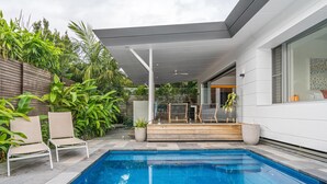 Un espace piscine extérieur moderne, doté d’une terrasse en bois, de deux chaises longues et d’un coin salon abrité.