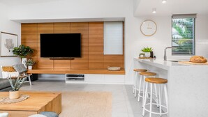 Un salon moderne doté d’un téléviseur à écran plat, d’un mur recouvert de panneaux en bois et d’un coin cuisine avec des tabourets de bar.
