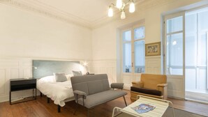 Quarto com cama, sofá cinza, mesa pequena, cadeira e janela com vista.