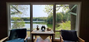A room with a large window offering a view of a lake and forest. There is a table with a vase, a bowl, and a book. Two chairs are positioned in front of the window. - Fiebing's Rustic/Remote Cabin on two Private Lakes (Traverse City)