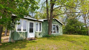 A small, single-story house with a green exterior and white trim, surrounded by trees and a grassy yard.