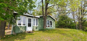 A small, single-story house with a green exterior and white trim, surrounded by trees and a grassy yard. - Fiebing's Rustic/Remote Cabin on two Private Lakes (Traverse City)