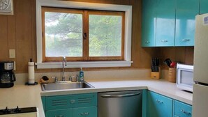 A kitchen with teal cabinets, a stainless steel sink, a window with a view of trees, and a microwave.