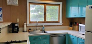 A kitchen with teal cabinets, a stainless steel sink, a window with a view of trees, and a microwave. - Fiebing's Rustic/Remote Cabin on two Private Lakes (Traverse City)