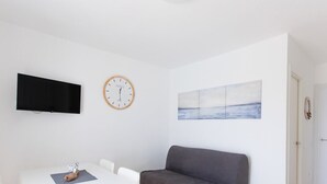 A minimalist room with a flat-screen TV, a wooden-framed clock, a painting, a gray sofa, and a white door.