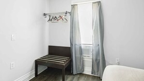 A small, sparsely furnished room with a bed, a bench, a coat rack, and a window with curtains. - Downtown Detroit 2BR | King, WiFi, Parking (Detroit)