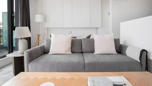 Een moderne woonkamer met een grijze bank, witte kussens, een bijzettafeltje met een lamp en een houten salontafel.