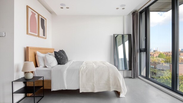 Een moderne slaapkamer met een groot raam, een bed met wit beddengoed, een nachtkastje met een lamp en lijstwerk op de muur.