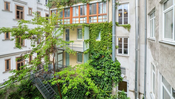 A multi-story building with a balcony on the second floor, surrounded by greenery and a metal staircase.