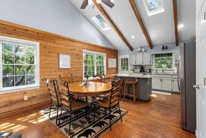 Une cuisine spacieuse avec des poutres en bois, un coin repas avec une table ronde et des chaises, et une vue sur l’extérieur grâce à de grandes fenêtres.