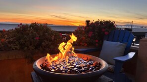 A fire pit with rocks and glass pebbles, a blue chair, and a view of the sunset over the water.