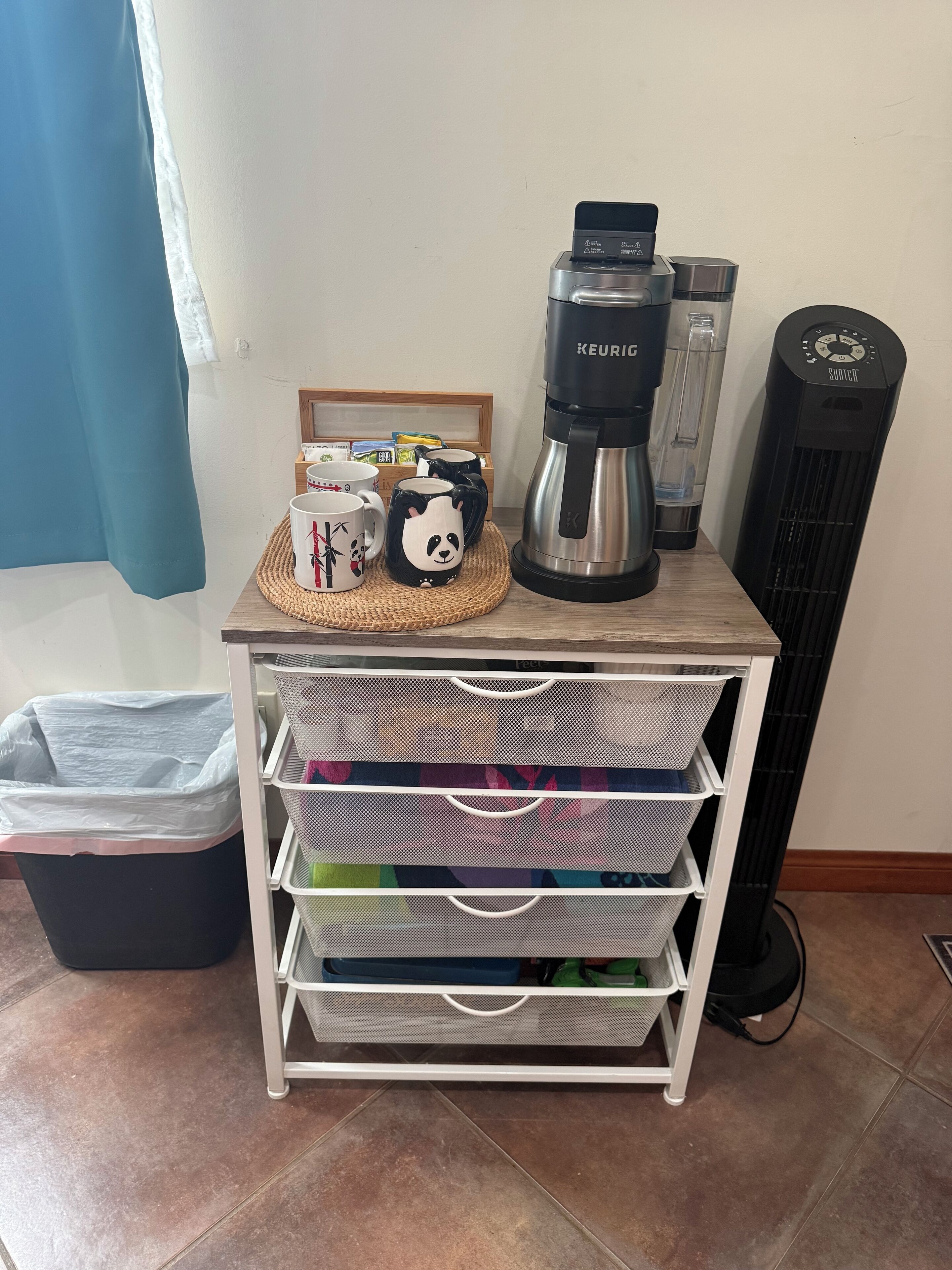 Una kitchenette con cafetera, dispensador de agua y calentador portátil. Se observan cestas de almacenamiento y un cubo de basura.