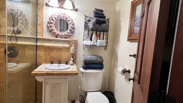 A bathroom with a sink, toilet, and shower. There is a towel rack with folded towels and a toilet paper holder.