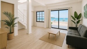 Een ruime woonkamer met een grote schuifpui naar uitzicht op het strand, een trap en een gezellig zitgedeelte met een bank en een kleine tafel.