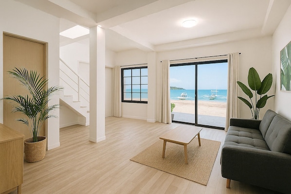 Amplia sala de estar con un gran ventanal corredera que da a una vista de la playa, una escalera y un rincón de descanso acogedor con un sofá y una mesita.