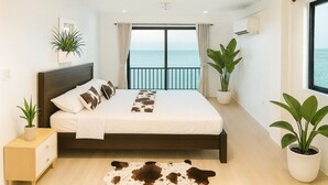 Een slaapkamer met een bed, nachtkastje, een kamerplant, een raam met uitzicht op zee en een airconditioner.