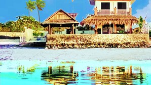 Een strandhuis met een hoofdgebouw en een kleinere constructie, beide met rieten daken, met uitzicht op een heldere blauwe zee.