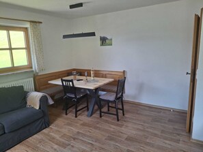 A room with a wooden floor, a table with chairs, a bench, a window with curtains, and a door.