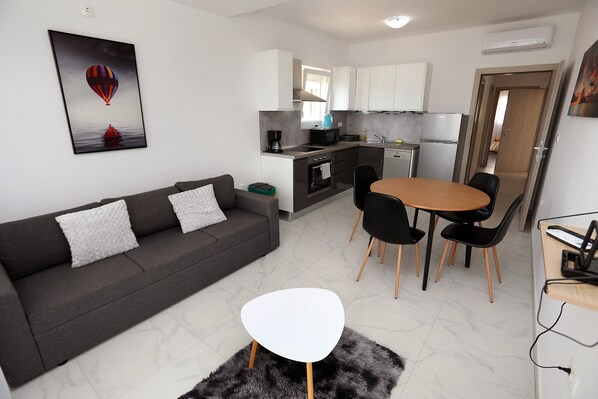 A modern living room with a grey sofa, a white coffee table, and a dining area with a round table and chairs. - Charming 2-Bed Apartment in Okrug Gornji (Okrug Gornji)