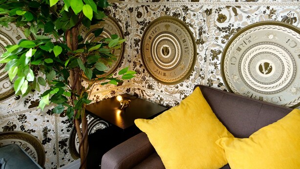 A room with a patterned wallpaper, a potted plant, a lamp, and a sofa with a yellow pillow. - Ultra Deluxe Versace Apartment Near Sheffield (Rotherham)