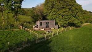 A small, modern cabin with a wooden exterior, situated on a grassy hill surrounded by trees. - 1 Bed Hut - Sleeps 4 - Pets - Parking (Minehead)