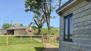 A rustic wooden cabin with a large window, surrounded by a fenced yard and a tree. - 1 Bed Hut - Sleeps 4 - Pets - Parking (Minehead)