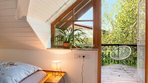 Ein Schlafzimmer mit einem Bett, einem Nachttisch mit Lampe, einem Fenster und Blick auf einen Balkon und Bäume.