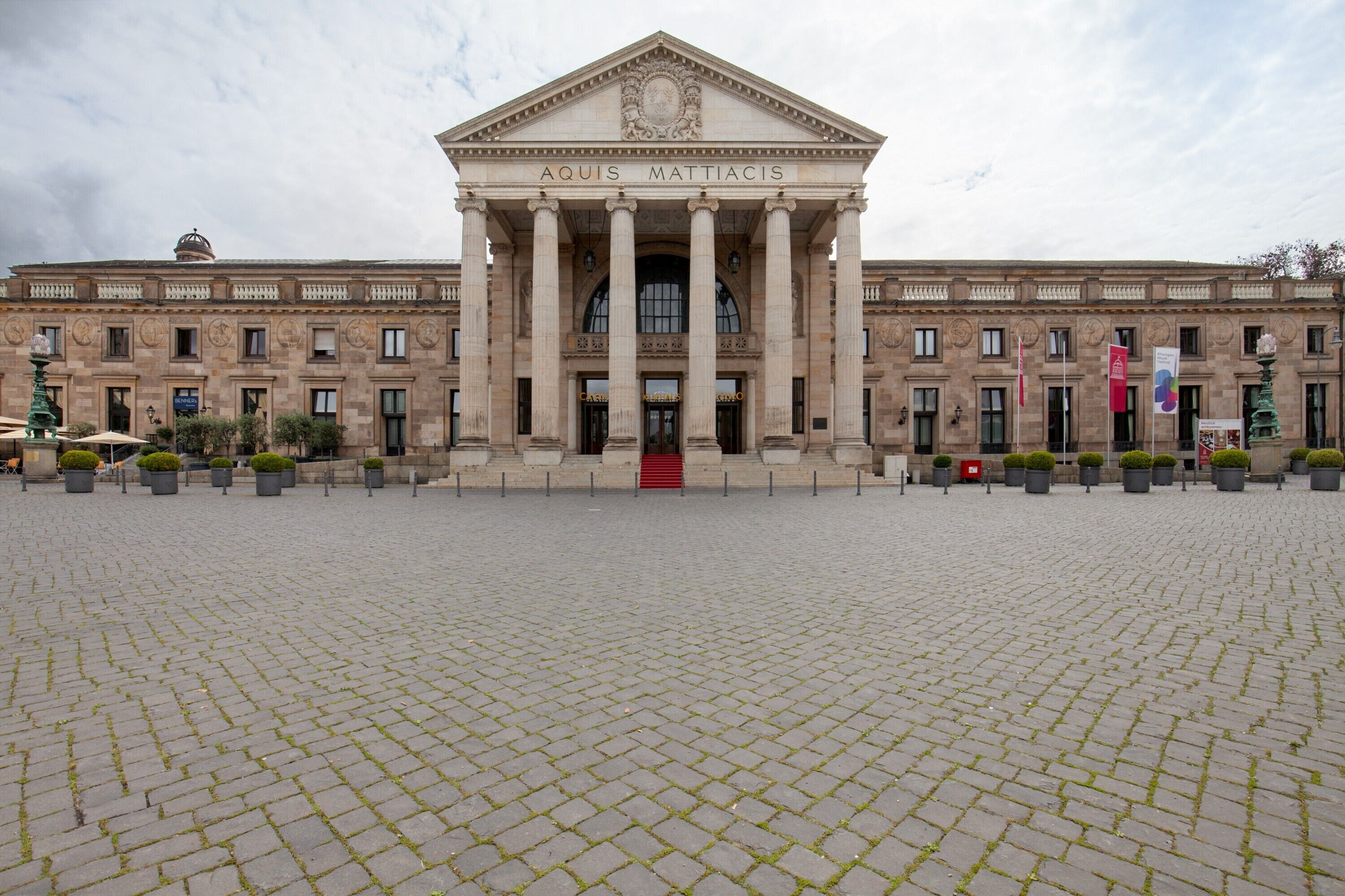 Ein großes Gebäude mit einem zentralen Eingang, der von Säulen gesäumt wird, einer roten Treppe und einem gepflasterten Innenhof.