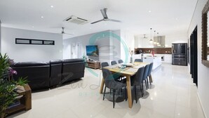 A modern living room with a black leather sofa, a dining table with chairs, and a kitchen area in the background. - ZENHOUSE - 5BR Family with Private Pool (Johnston)