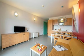 Un salon moderne doté d’un téléviseur à écran plat, de meubles en bois, d’un coin repas avec une table et des chaises, et d’une cuisine avec des armoires en bois et un luminaire suspendu.