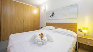 Un lit bien fait, avec du linge de lit et des serviettes blanches, une armoire en bois, une table de chevet avec une lampe et un tableau accroché au mur.