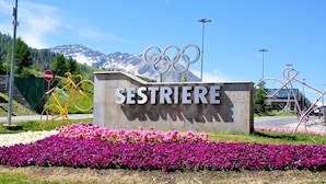 Une pancarte portant l’inscription « SESTRIERE » et les anneaux olympiques, entourée de fleurs et avec une chaîne de montagnes en arrière-plan.