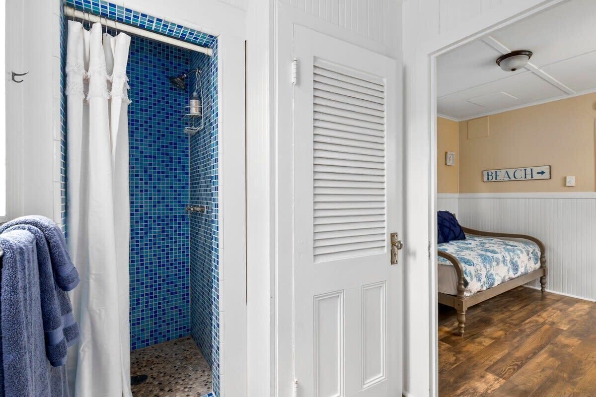 A bathroom with a shower, a door leading to a bedroom with a bed and a sign indicating a beach direction.
