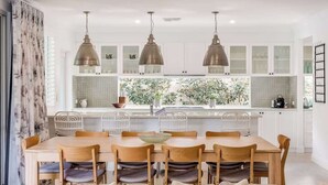 A modern kitchen with white cabinets, a dining table with chairs, and pendant lights. - Hamptons By The Green—Grand Golfside Retreat (DUNSBOROUGH)