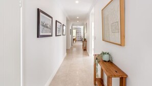 A hallway with framed pictures on the walls, a wooden bench with a plant, and a tiled floor. - Hamptons By The Green—Grand Golfside Retreat (DUNSBOROUGH)