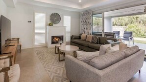 A modern living room with a fireplace, a sofa set, a television, and a dining area. - Hamptons By The Green—Grand Golfside Retreat (DUNSBOROUGH)