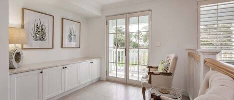 Una habitación amplia con un gran ventanal corredero que da a un balcón, un sillón mullido, una pequeña mesa redonda y un armario con una lámpara.