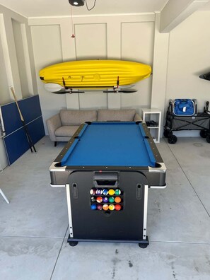 A pool table with a blue felt surface, a yellow paddleboard, a pool cue, and a blue bag in a room with a gray couch and a white cabinet.