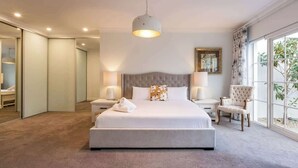 A hotel bedroom with a large bed, bedside lamps, a chair, and a view through the window. - Hamptons By The Green—Grand Golfside Retreat (DUNSBOROUGH)