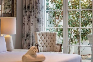 A bedroom with a bed, a tufted armchair, a window with a view of greenery, and a lamp. - Hamptons By The Green—Grand Golfside Retreat (DUNSBOROUGH)