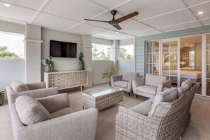 A spacious living area with a ceiling fan, comfortable seating, a flat-screen TV, and a view through glass doors to a bedroom. - Hamptons By The Green—Grand Golfside Retreat (DUNSBOROUGH)