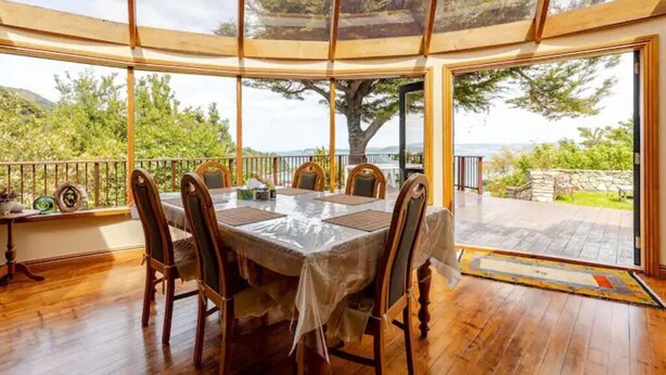 A dining area with a table set for six, surrounded by wooden chairs, with a view of trees and a stone wall. - Sea views from holiday house (Lower Hutt)