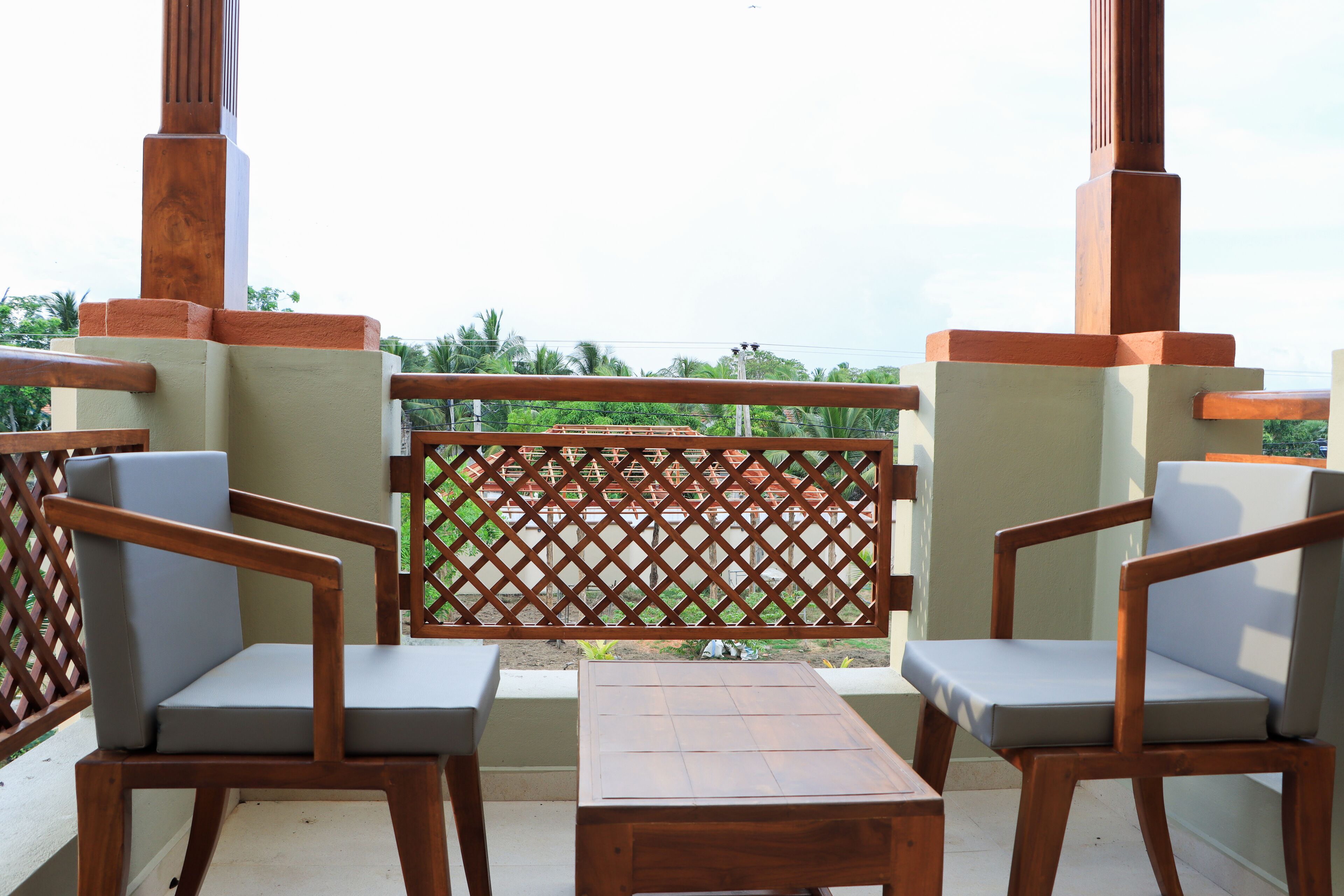 A balcony with two wooden chairs and a small table, a lattice partition, and a view of greenery.