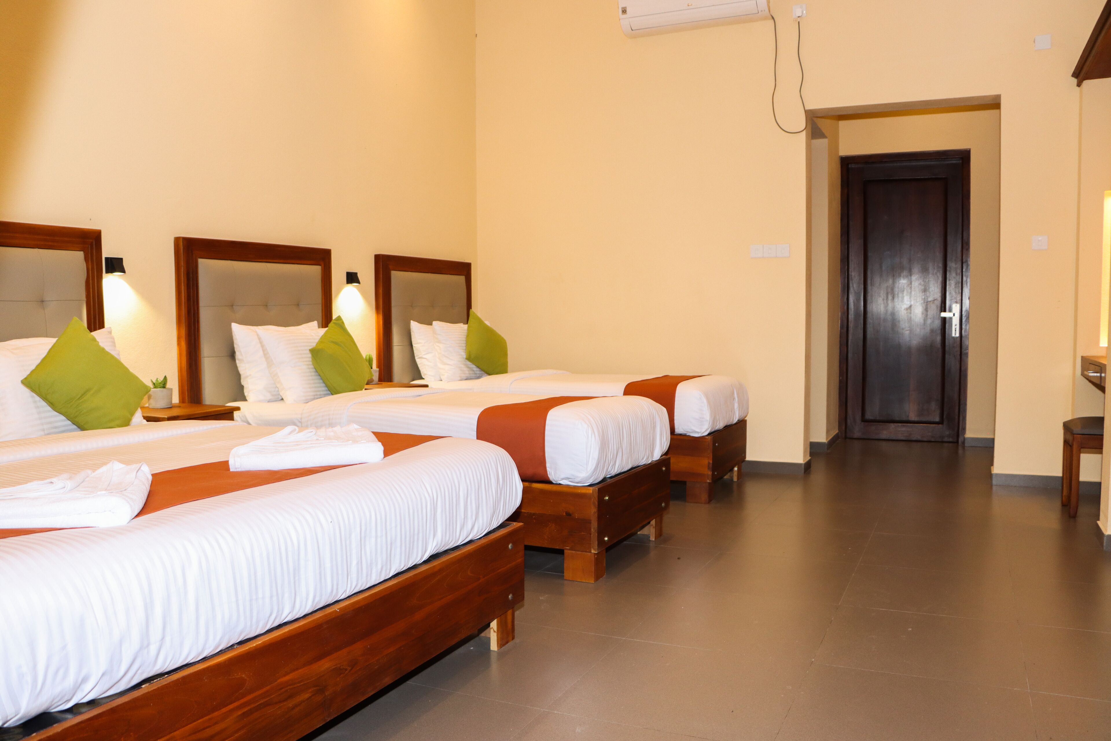 A hotel room with three wooden beds, each with white linens and green pillows. There is a wooden nightstand with a lamp, and a dark wooden door in the background.