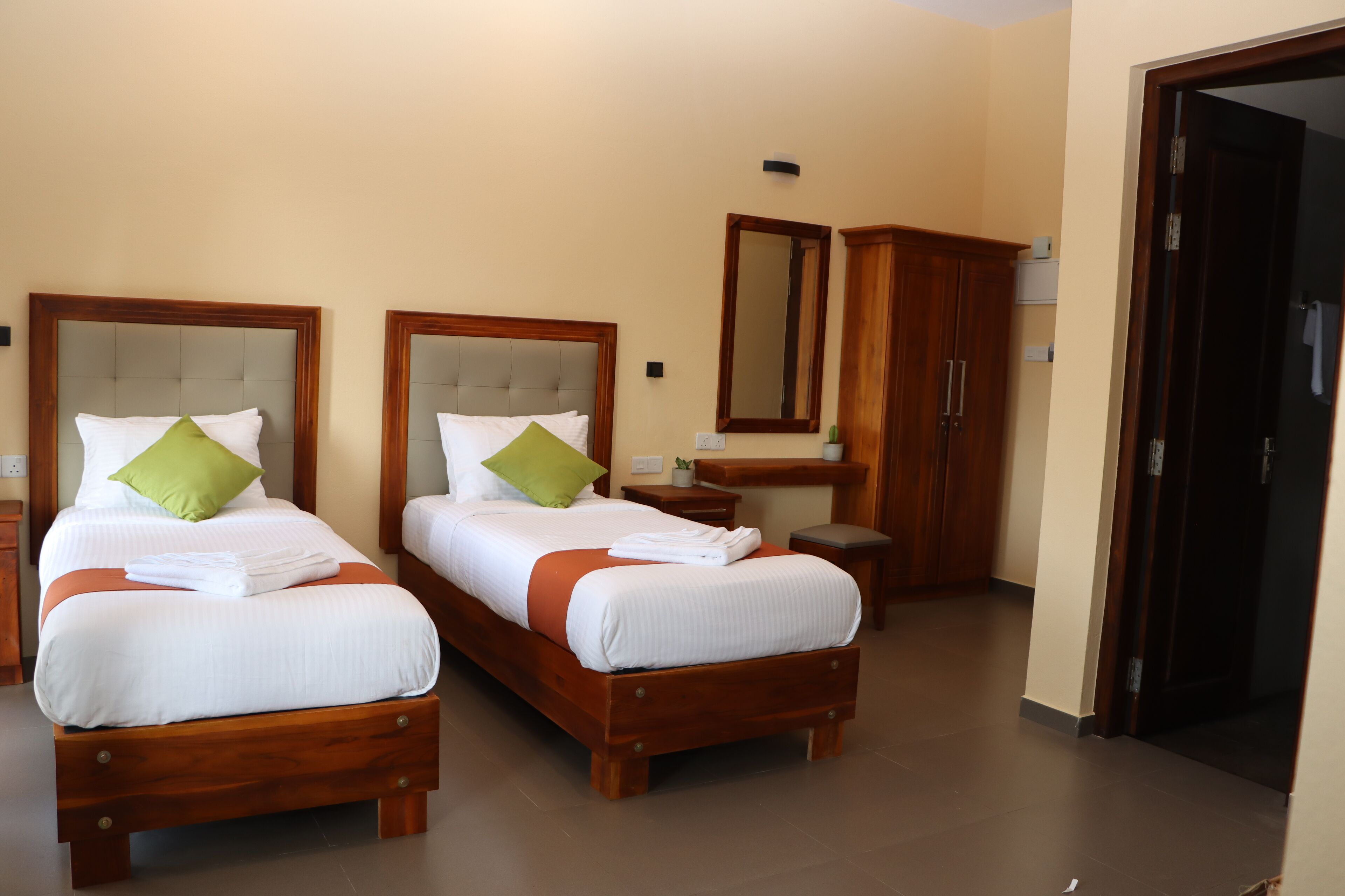 A hotel room with two beds, each with a wooden headboard, white bedding, and green pillows. There is a wooden wardrobe, a small table with a lamp, and a mirror on the wall.