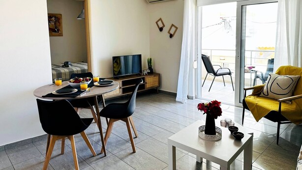 A modern living room with a dining area, a TV, and a balcony with chairs. - Cozy Snuggle- 2min To Beach, 6min Walk To Old Town (Chania)